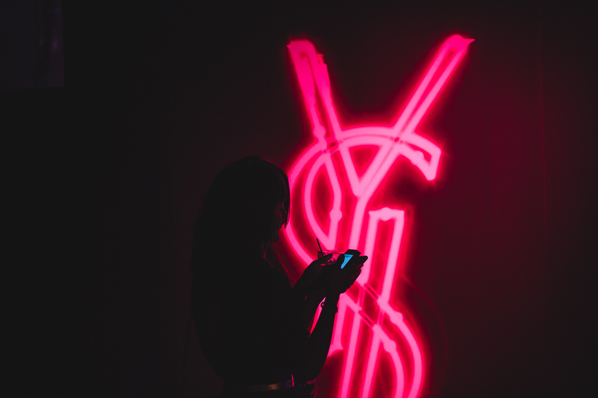 Silhouet van een vrouw met een drankje, staande voor een roze neon Yves Saint Laurent-logo.