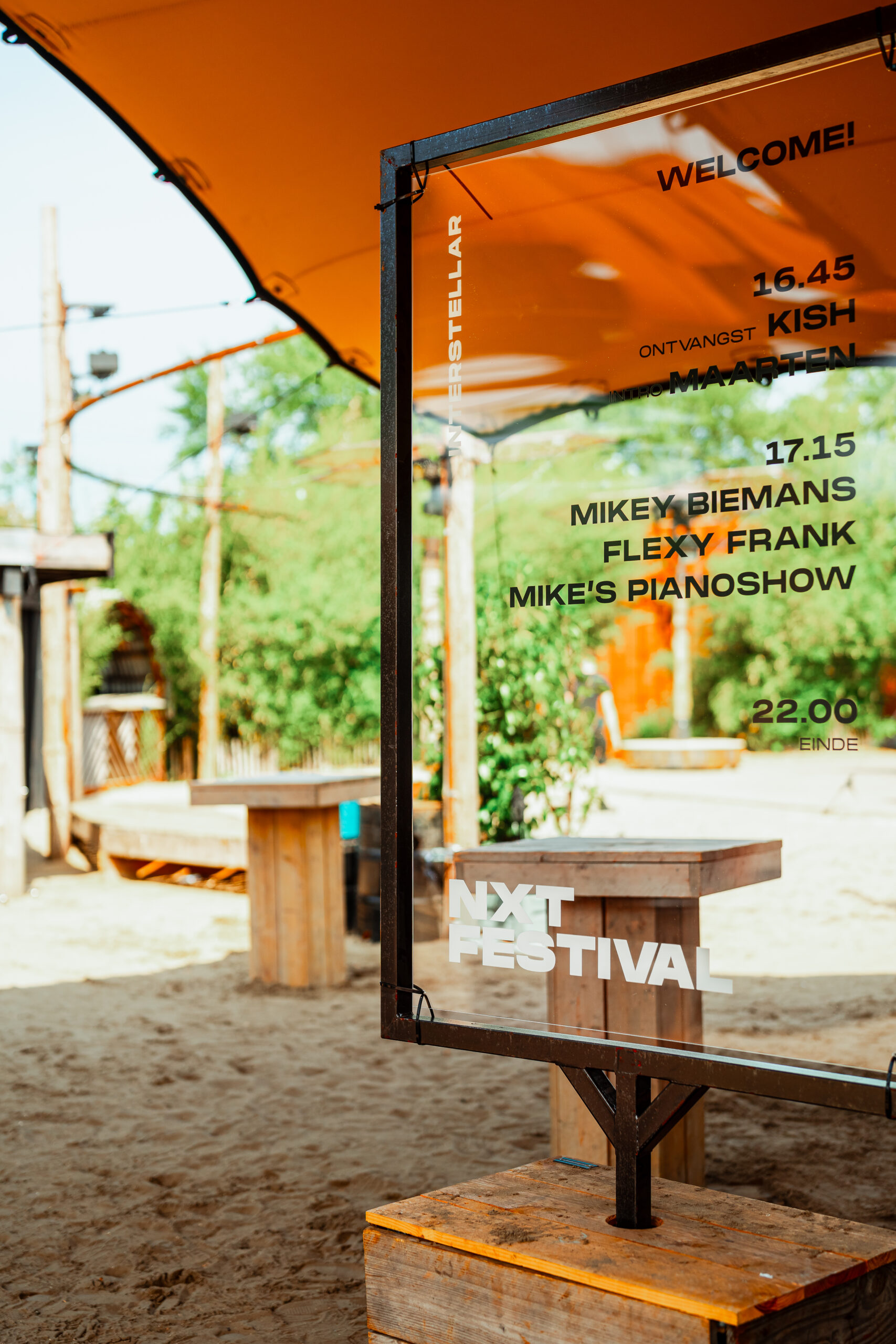Transparant festivalprogramma op metalen standaard onder een oranje tent.