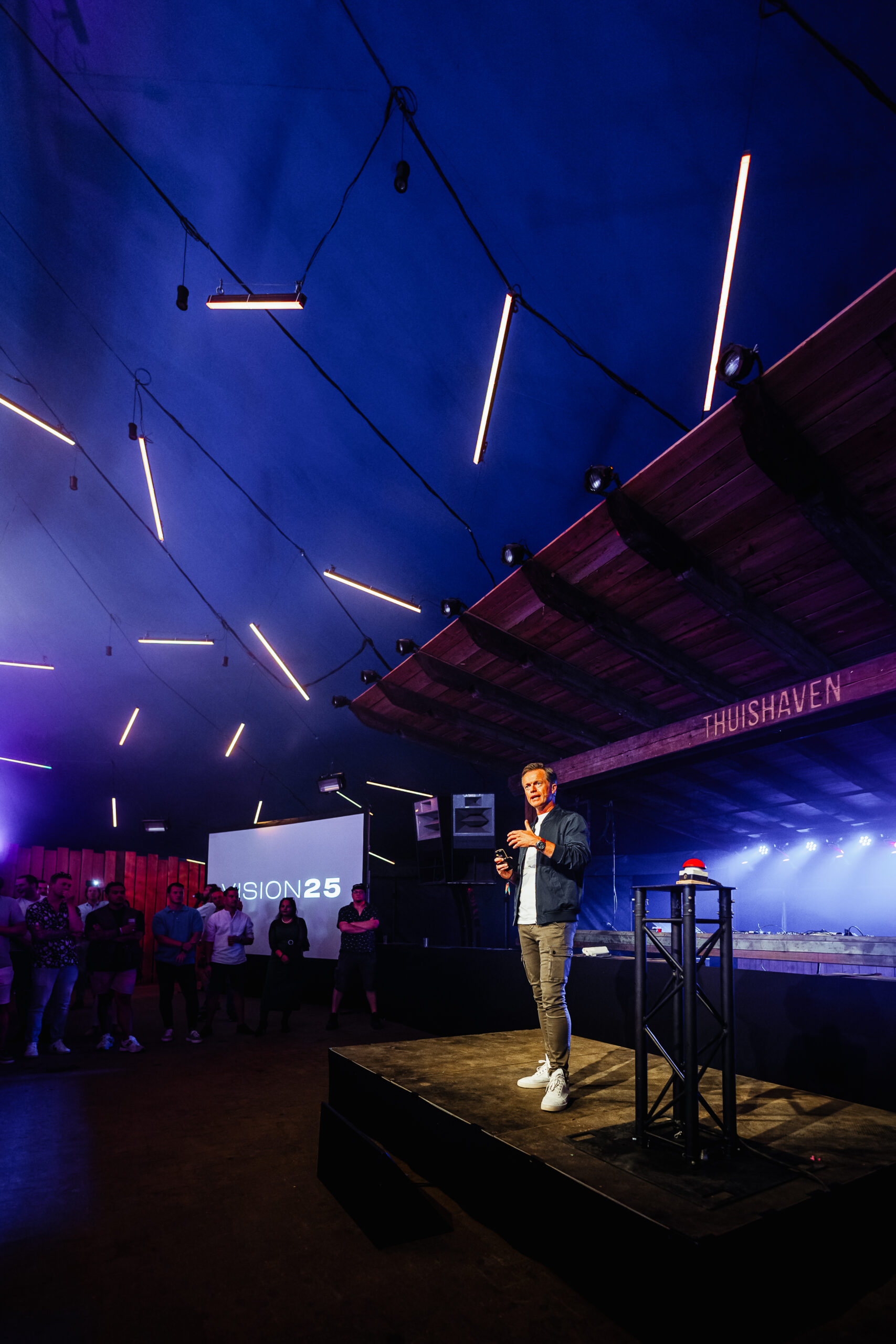 preker op podium in Thuishaven met blauwe sfeerverlichting en Vision 25 presentatie.
