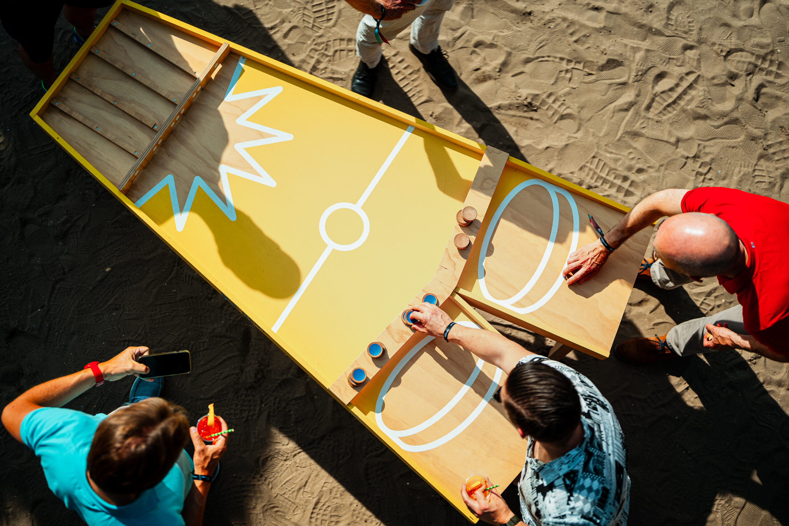 Geel houten sjoelspel op een zanderig festivalterrein.