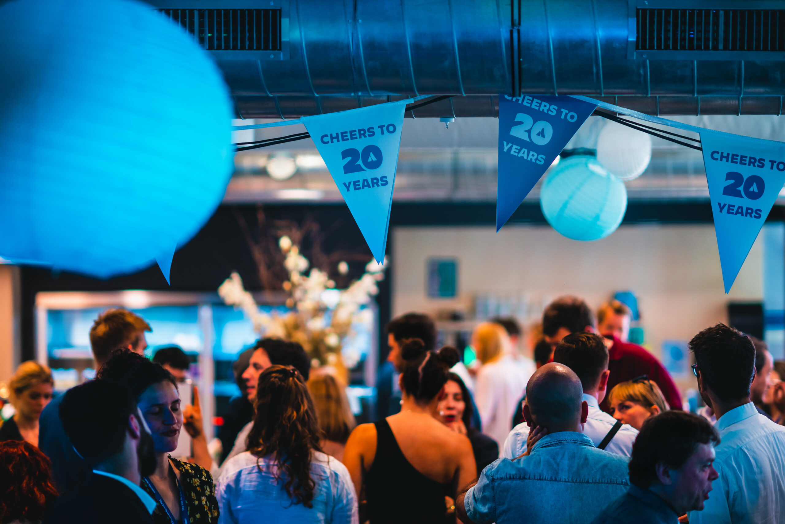 Groep mensen tijdens een jubileumevenement met feestelijke slingers waarop 'Cheers to 20 Years' staat.