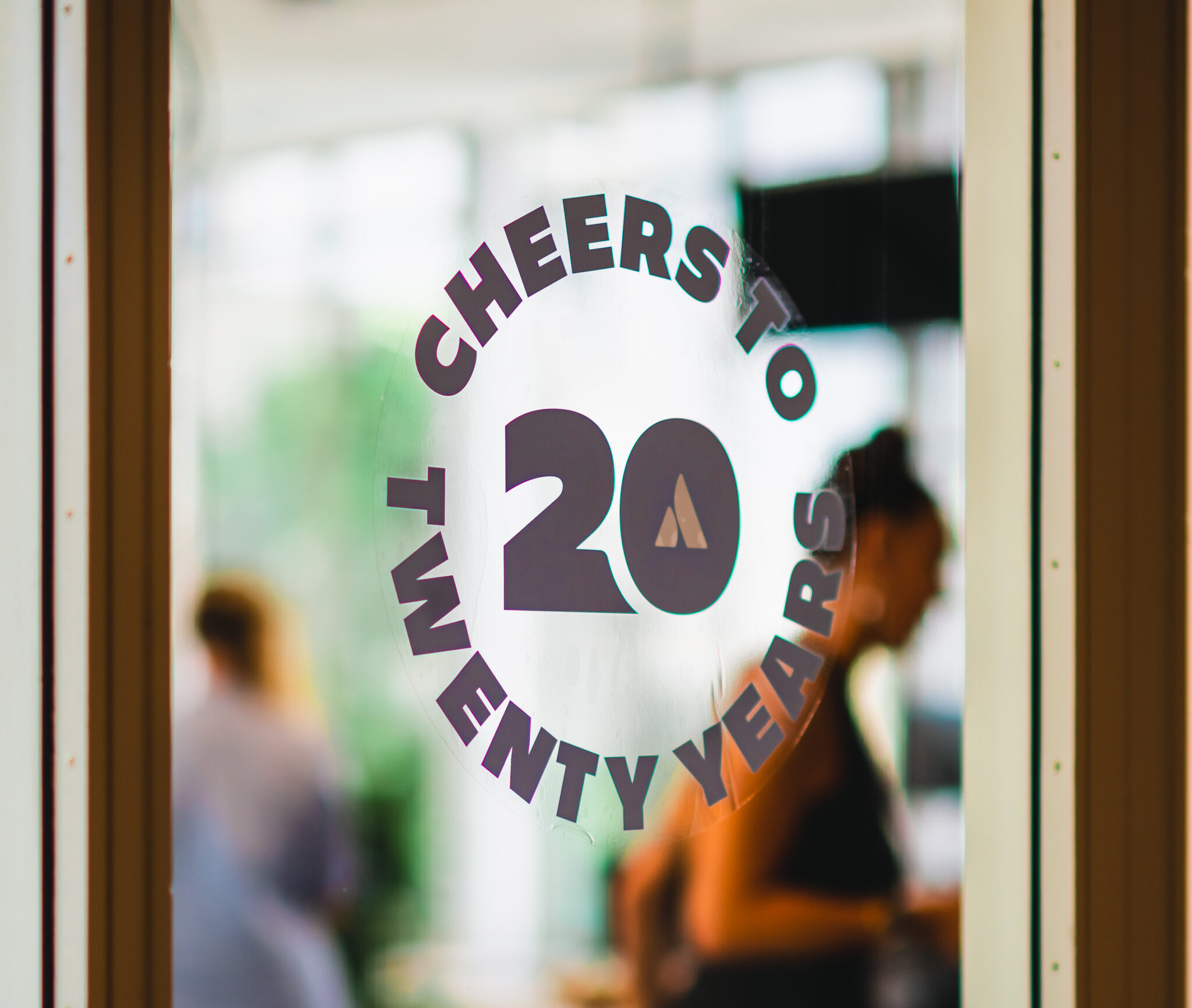 Jubileumsticker met de tekst 'Cheers to Twenty Years' op een glazen deur, met onscherpe achtergrond.