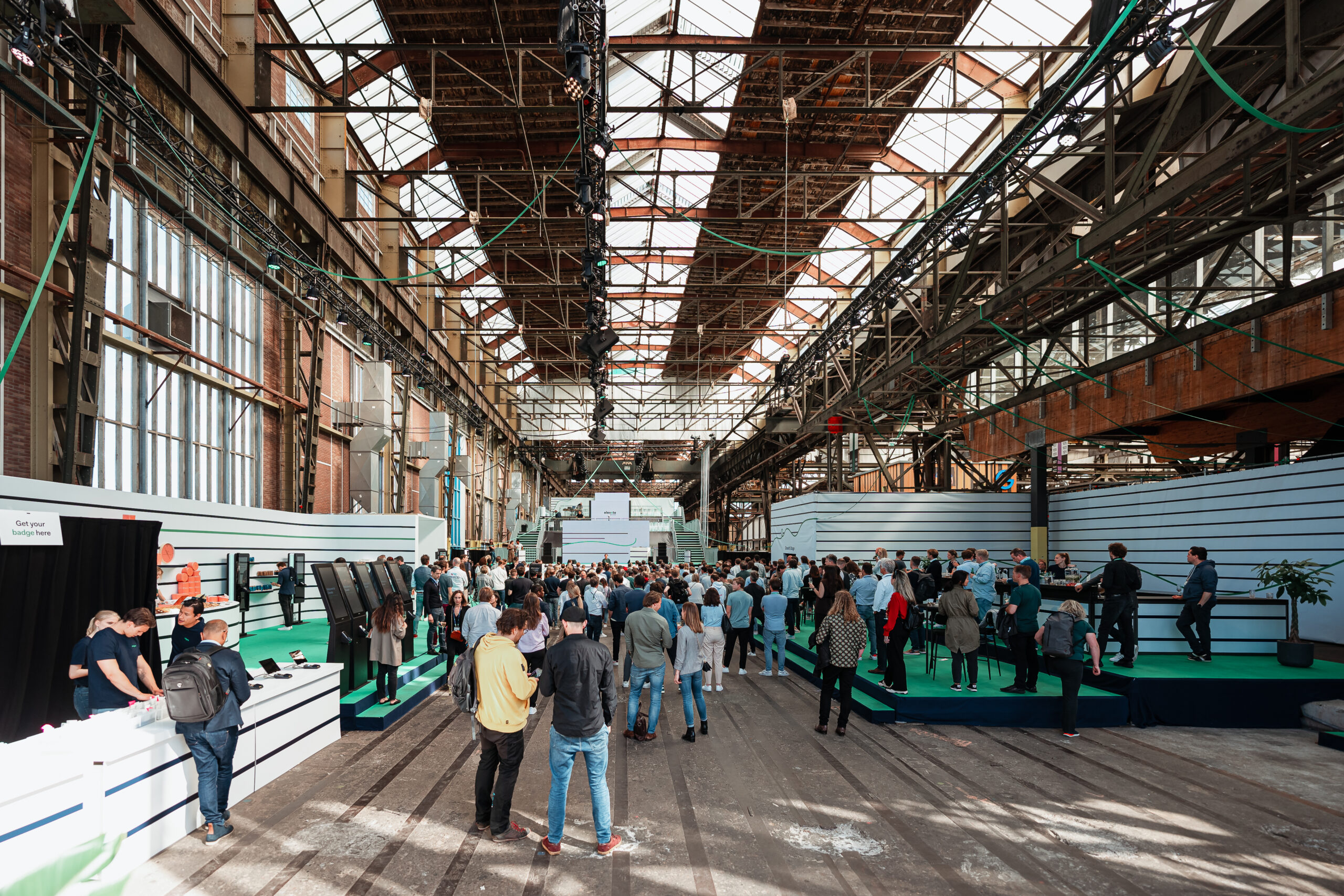 Klantendag organiseren - Een groot industrieel evenement in een voormalige fabriekshal met hoge glazen daken, waar een publiek samenkomt voor een presentatie en netwerkmogelijkheden.