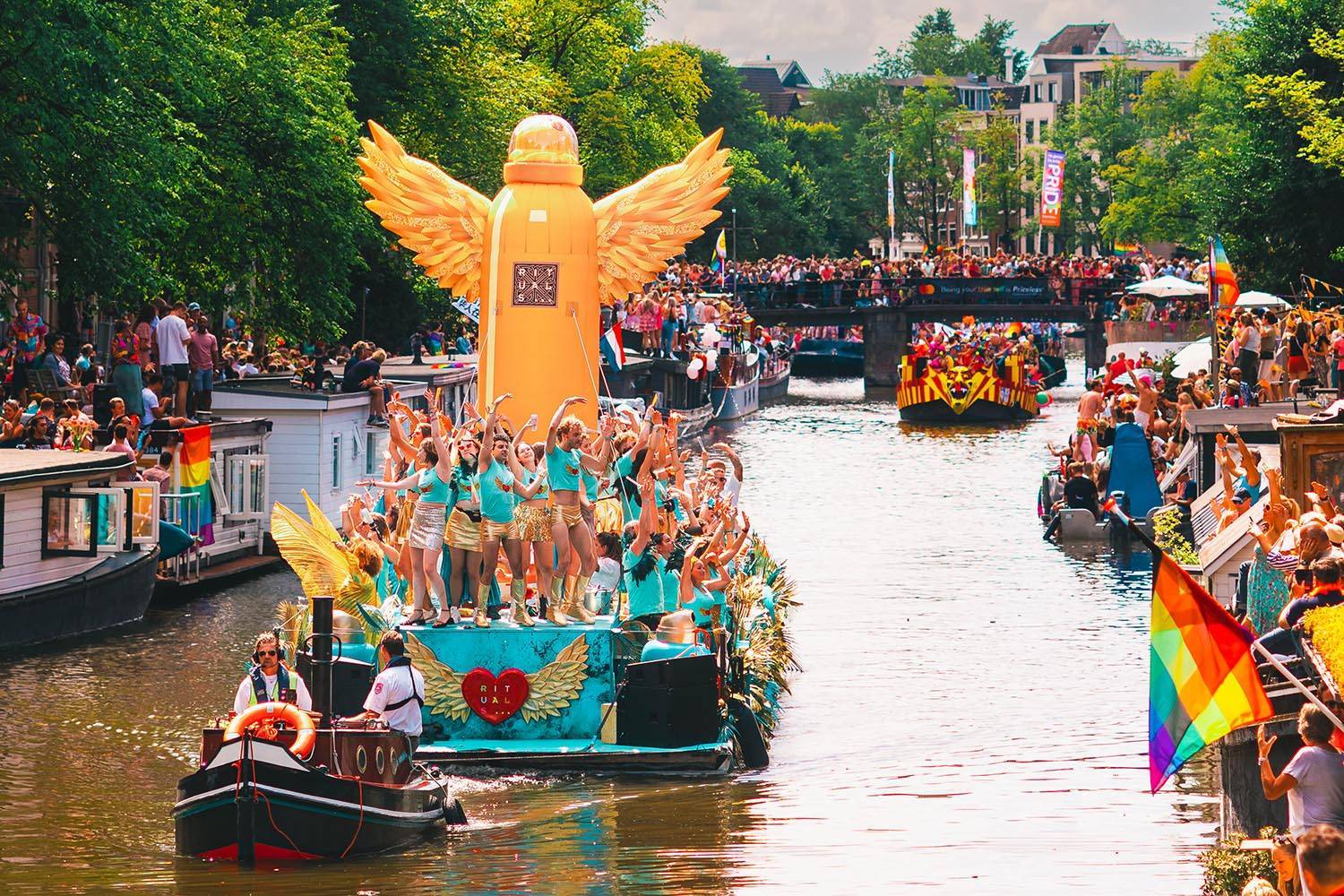 Vrolijke en kleurrijke botenparade tijdens Pride Amsterdam, met feestelijke deelnemers op een opvallende boot met gouden vleugels en regenboogvlaggen langs de grachten.