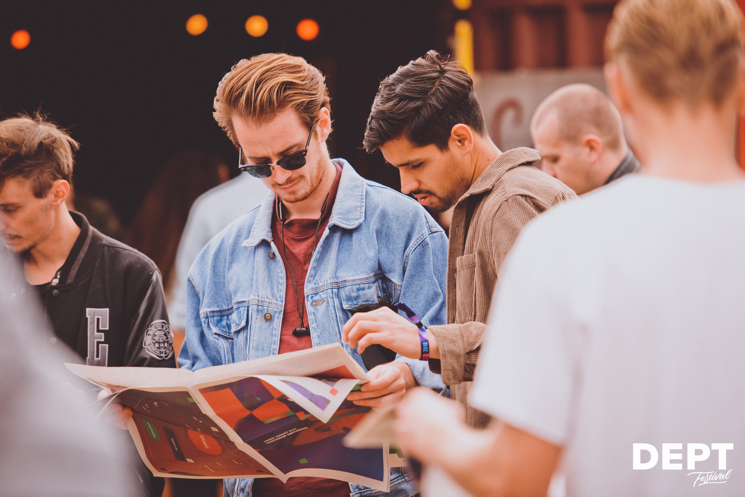 Relatie evenement - Twee jonge mannen bekijken een kleurrijke poster of festivalprogramma tijdens een buitenfestival, terwijl andere bezoekers op de achtergrond rondlopen.