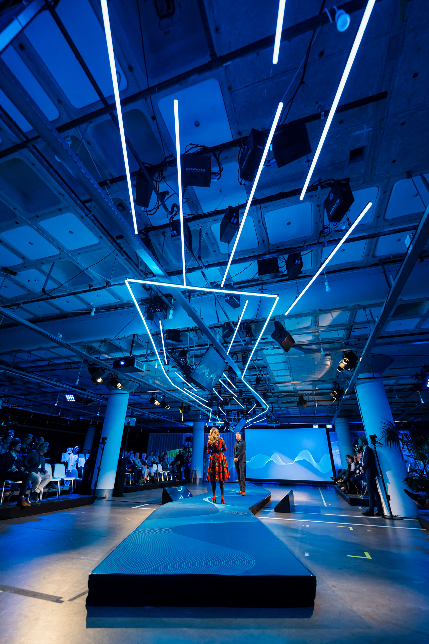 Futuristische blauwe verlichting en podiumontwerp tijdens het Statkraft 25-jarig jubileumevenement, met presentatrice op de achtergrond.