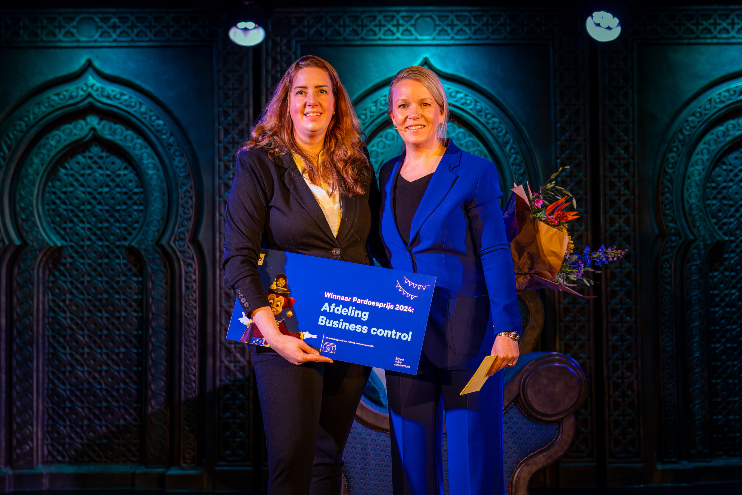 Twee vrouwen op een podium ontvangen een award, waarbij ze een blauw bord met de tekst "Winnaar Pardoesprijs 2024 - Afdeling Business Control" vasthouden.