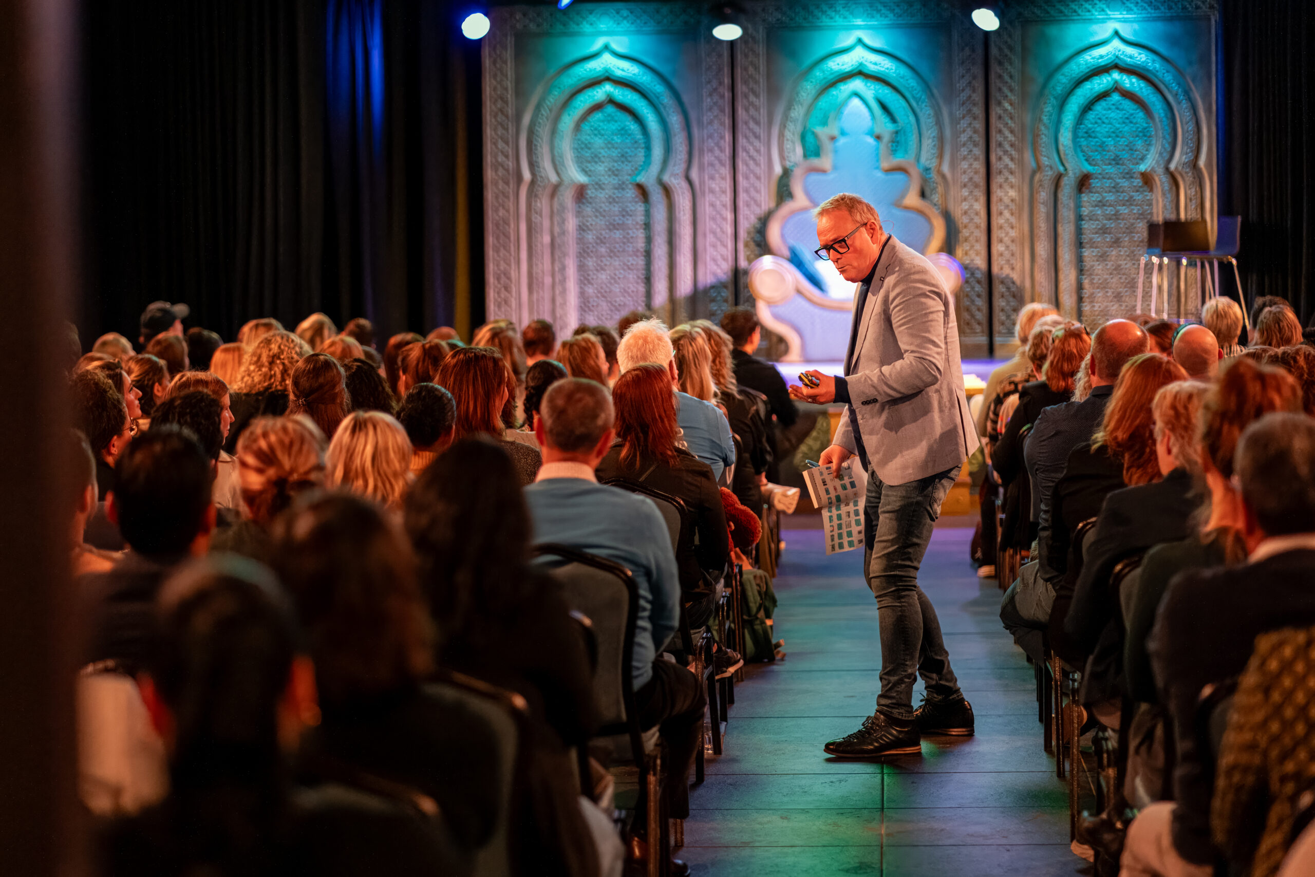 Een spreker in een lichtblauw colbert beweegt zich door het gangpad van een volle zaal tijdens een conferentie, met een sfeervol verlicht podium in de achtergrond.