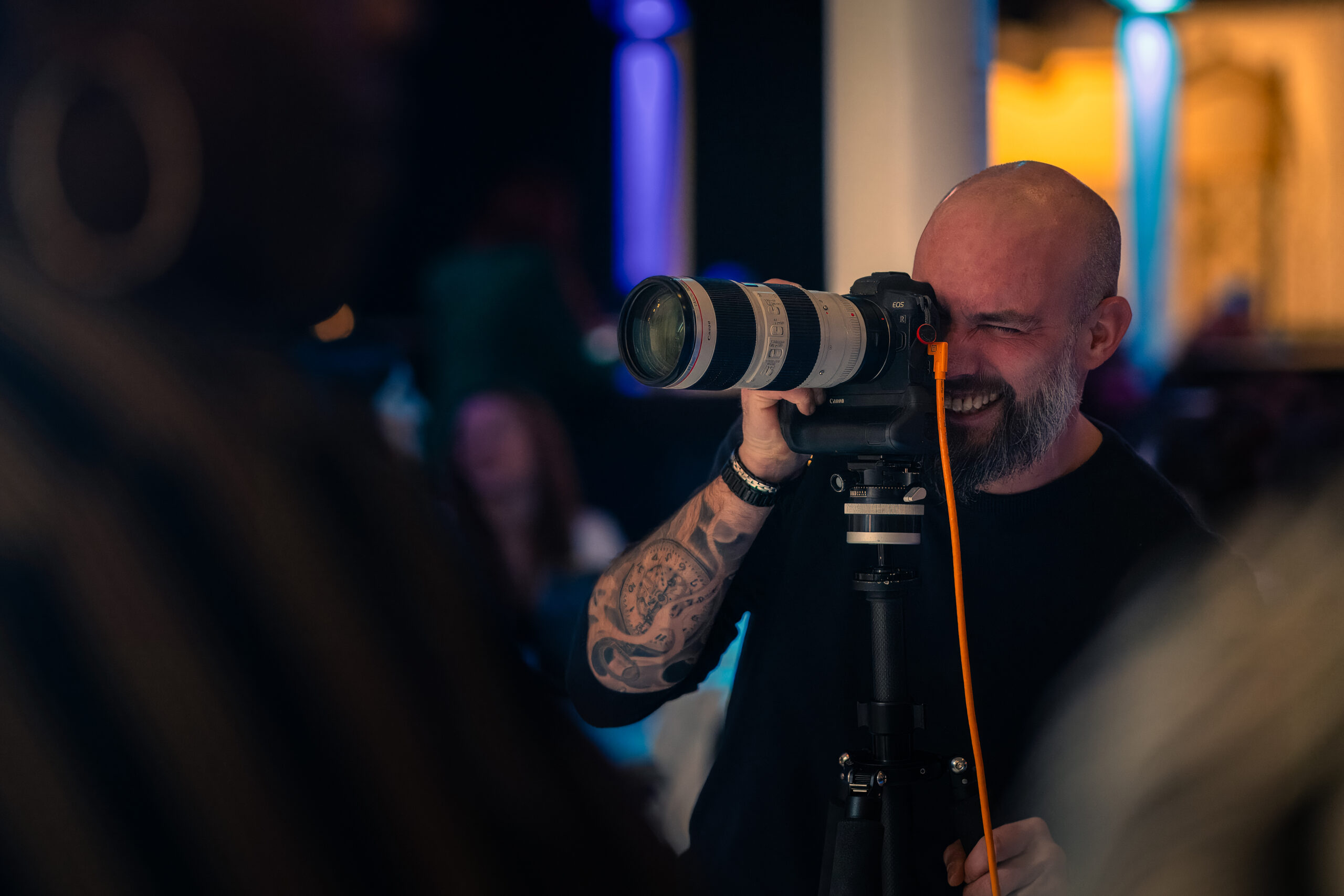 Een lachende fotograaf met een getatoeëerde arm richt zijn camera met telelens op een onderwerp, terwijl hij door de zoeker kijkt.