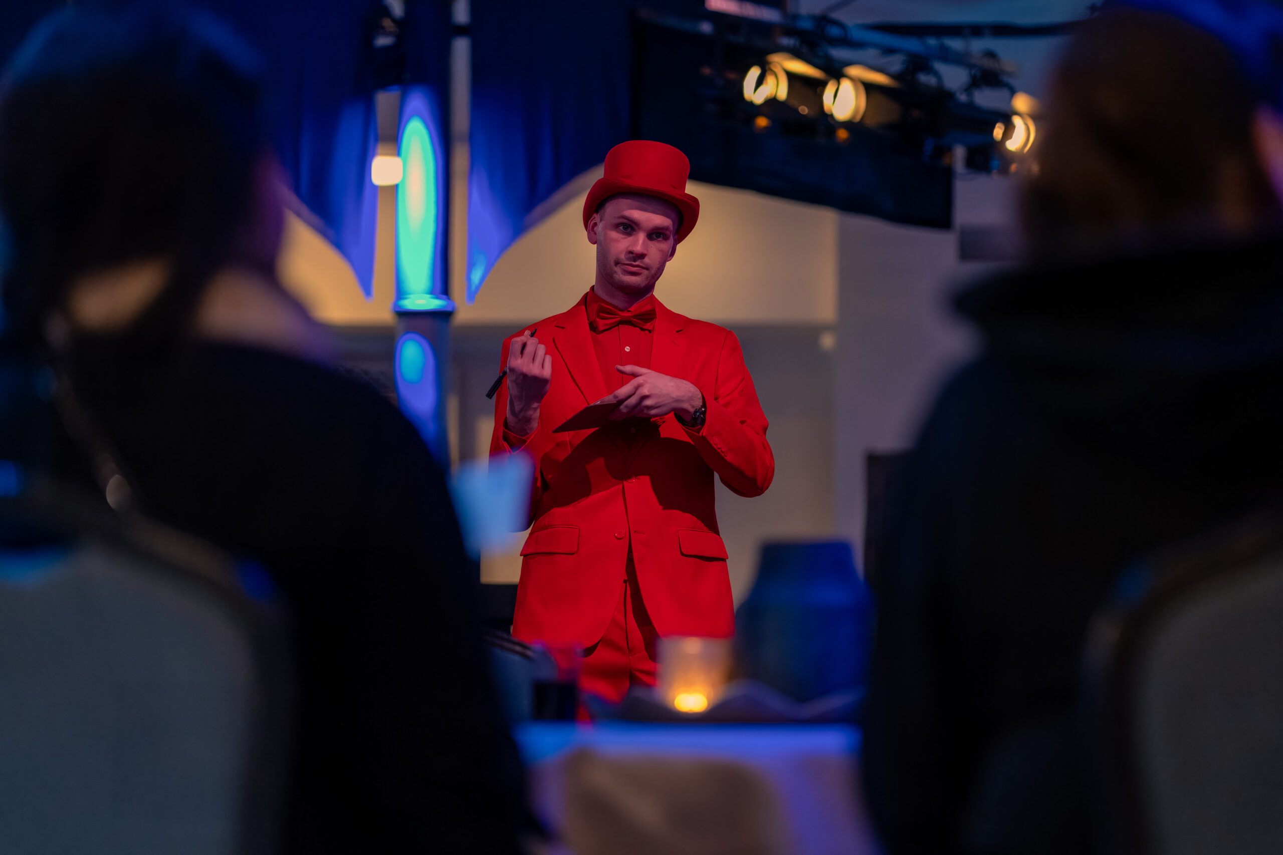 Een entertainer in een felrood pak met hoge hoed en strikje voert een act uit tijdens een evenement, terwijl het publiek aandachtig toekijkt.