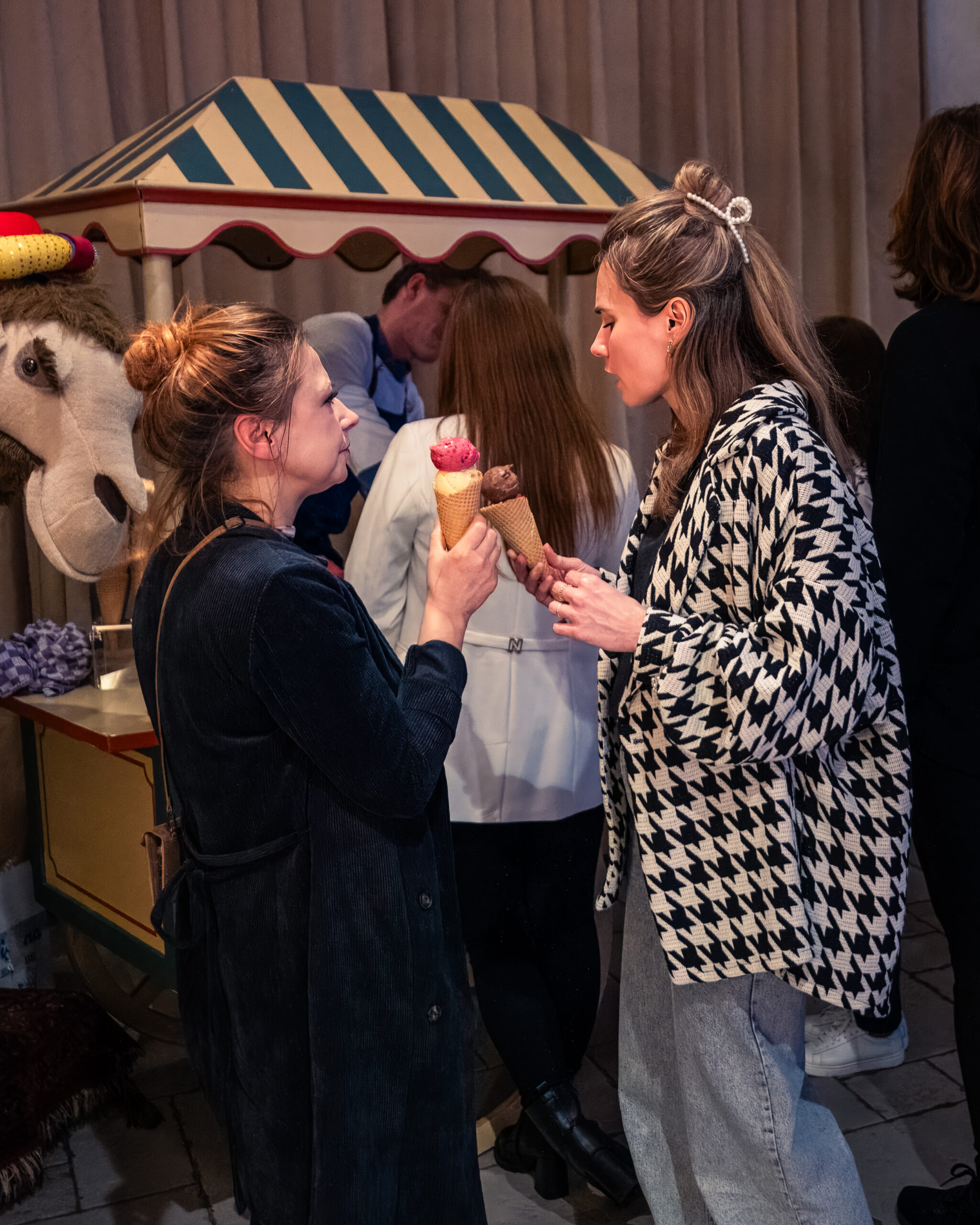 Twee vrouwen genieten van een ijsje terwijl ze in gesprek zijn bij een nostalgische ijscokar met blauw-witte strepen.