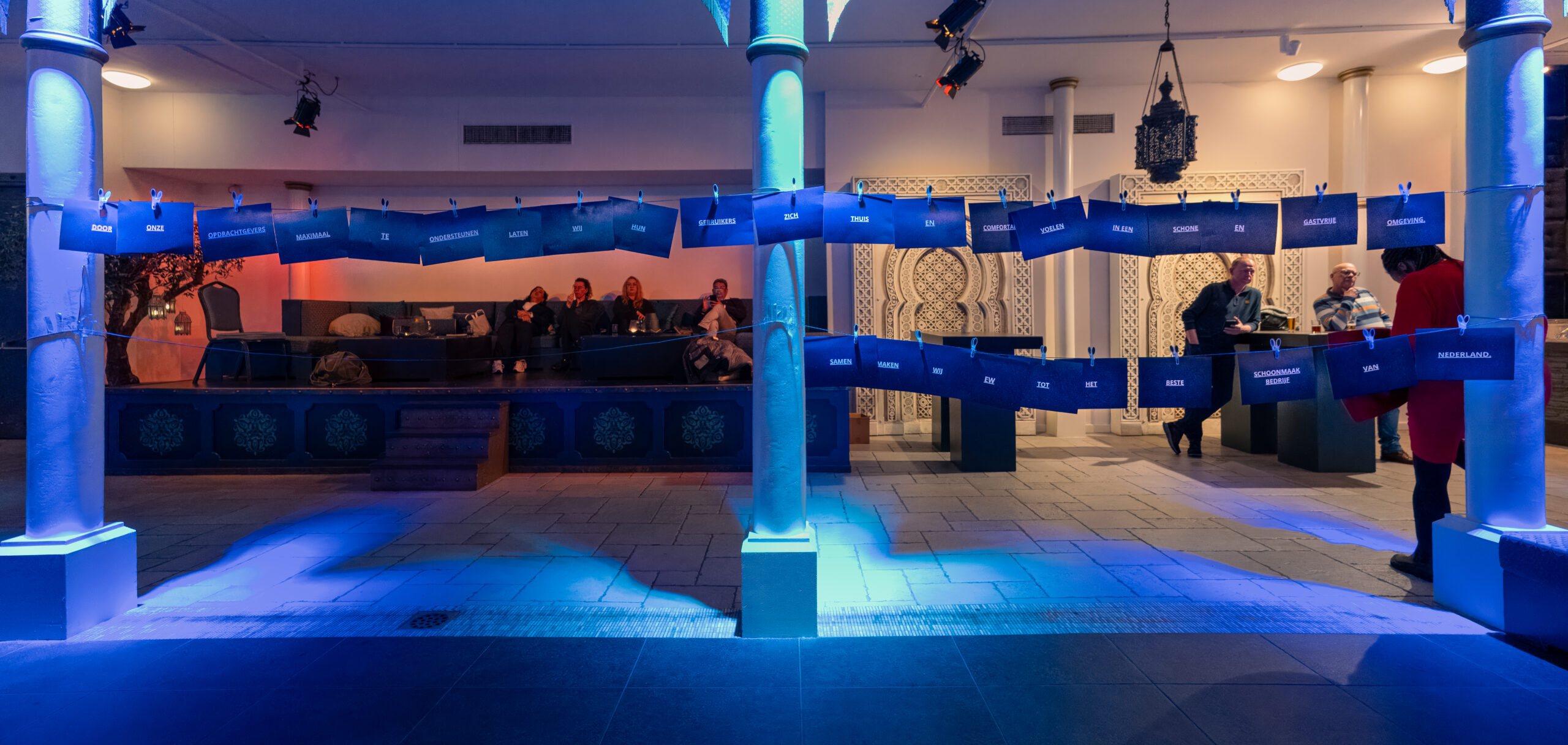 Blauwe kaarten met tekst hangen aan touwen tussen pilaren in een sfeervol verlichte ruimte met Oosterse decoratie en loungestoelen op de achtergrond.