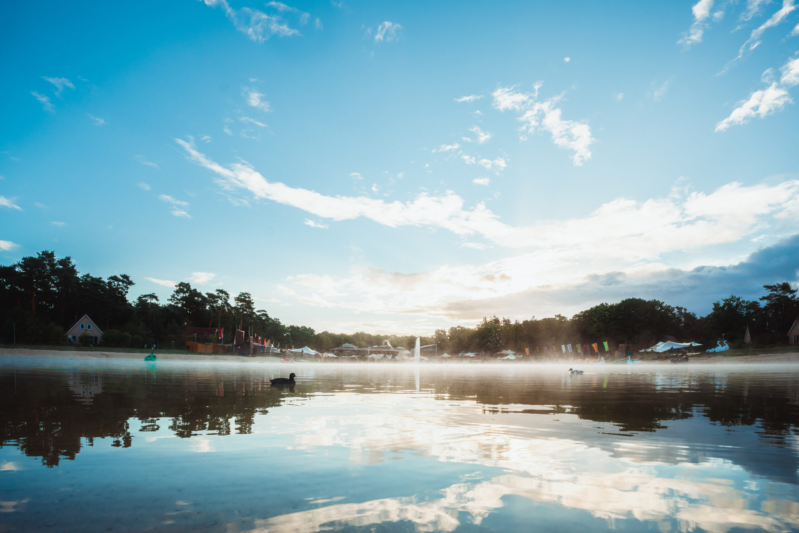 Een serene waterplas met een reflectie van de lucht, omringd door bomen en een festivalterrein met kleurrijke vlaggen en tenten.