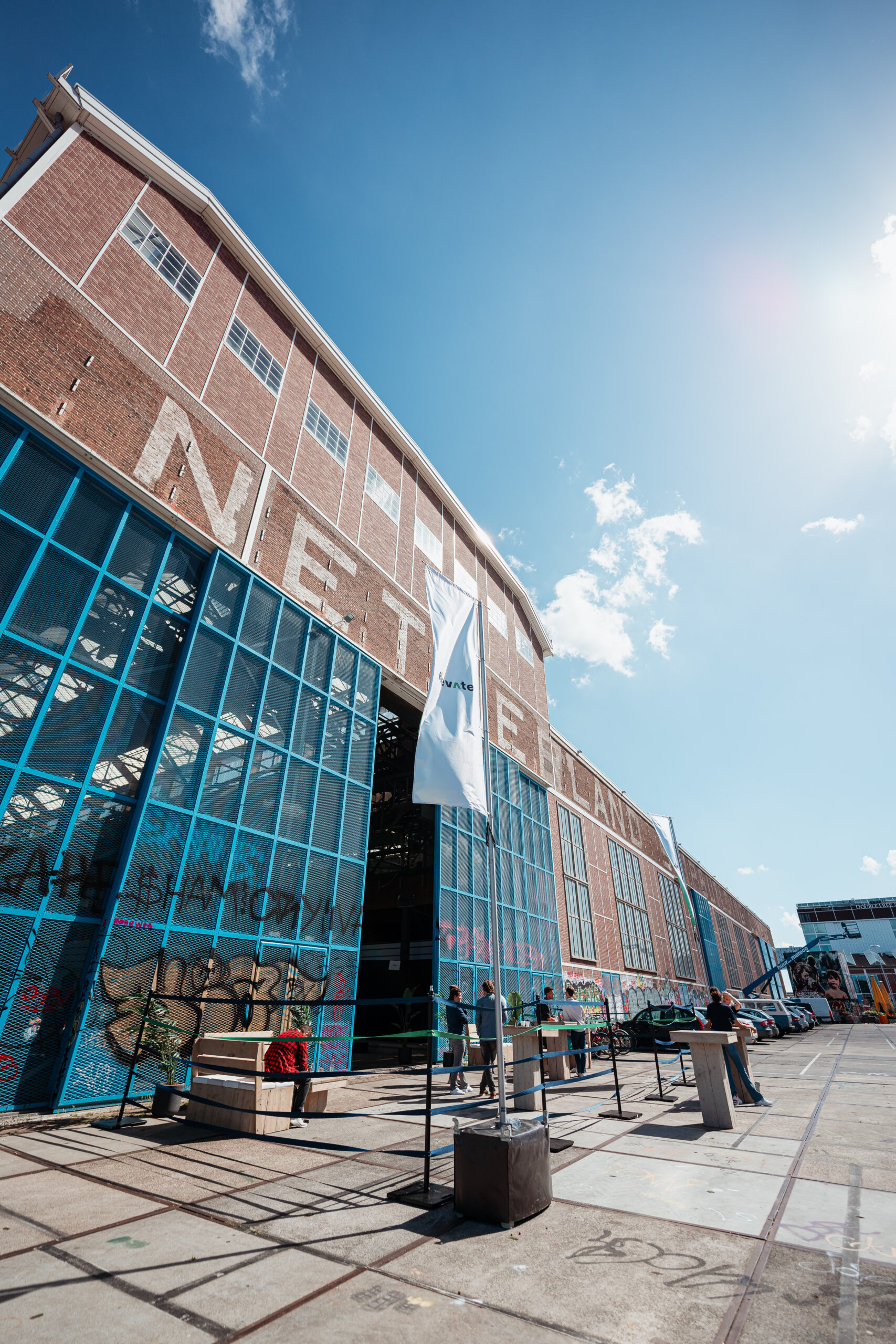 Industriële evenementenlocatie met een groot bakstenen gebouw met blauwe deuren en witte vlaggen, waar mensen zich verzamelen bij statafels onder een zonnige hemel.