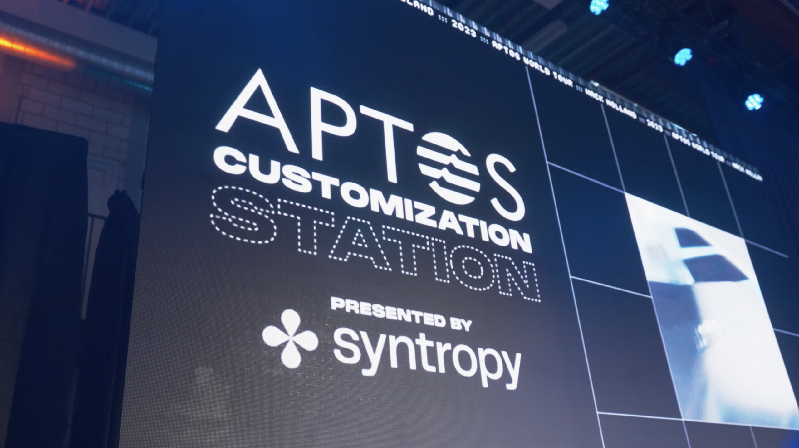 Een projectiescherm met de titel "Aptos Customization Station, presented by Syntropy.