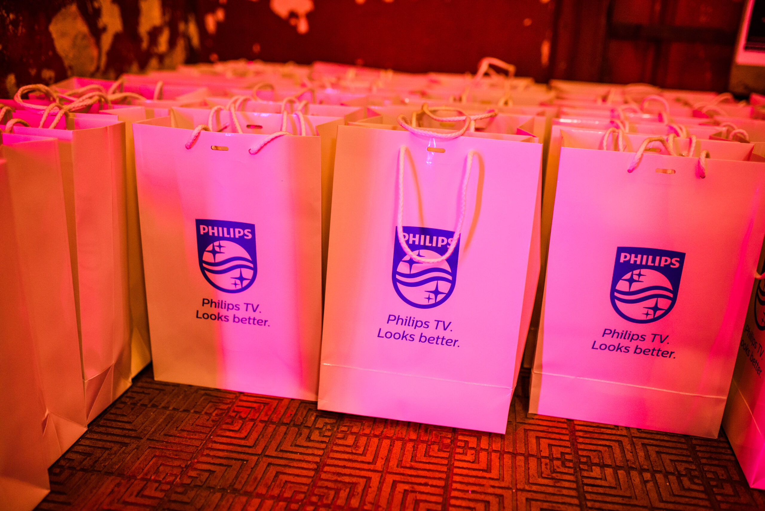Rij witte goodiebags met het Philips-logo en de tekst 'Philips TV. Looks better.' verlicht met roze tinten.