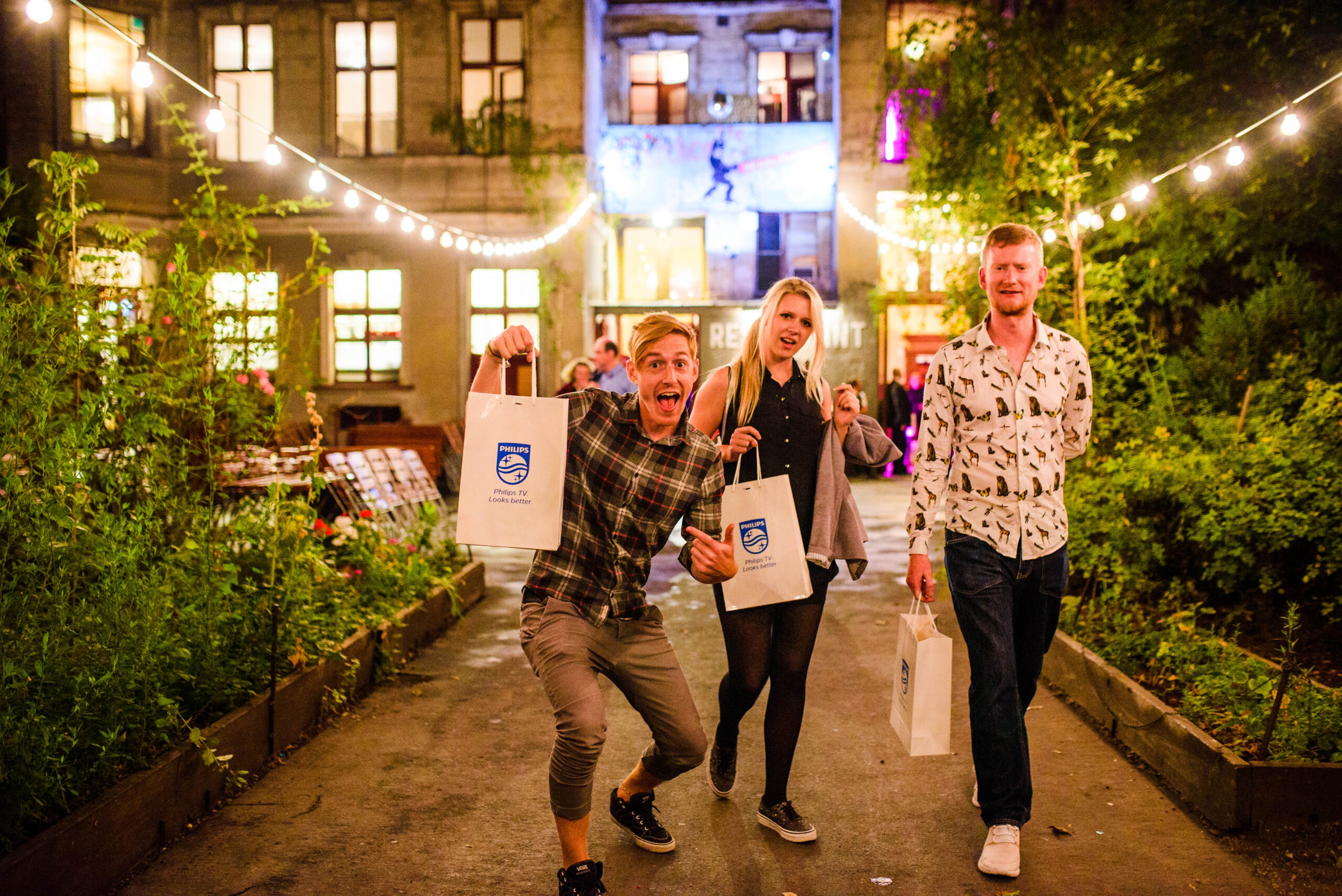 Drie blije gasten lopen buiten met goodiebags van Philips TV onder sfeervolle lichtslingers.
