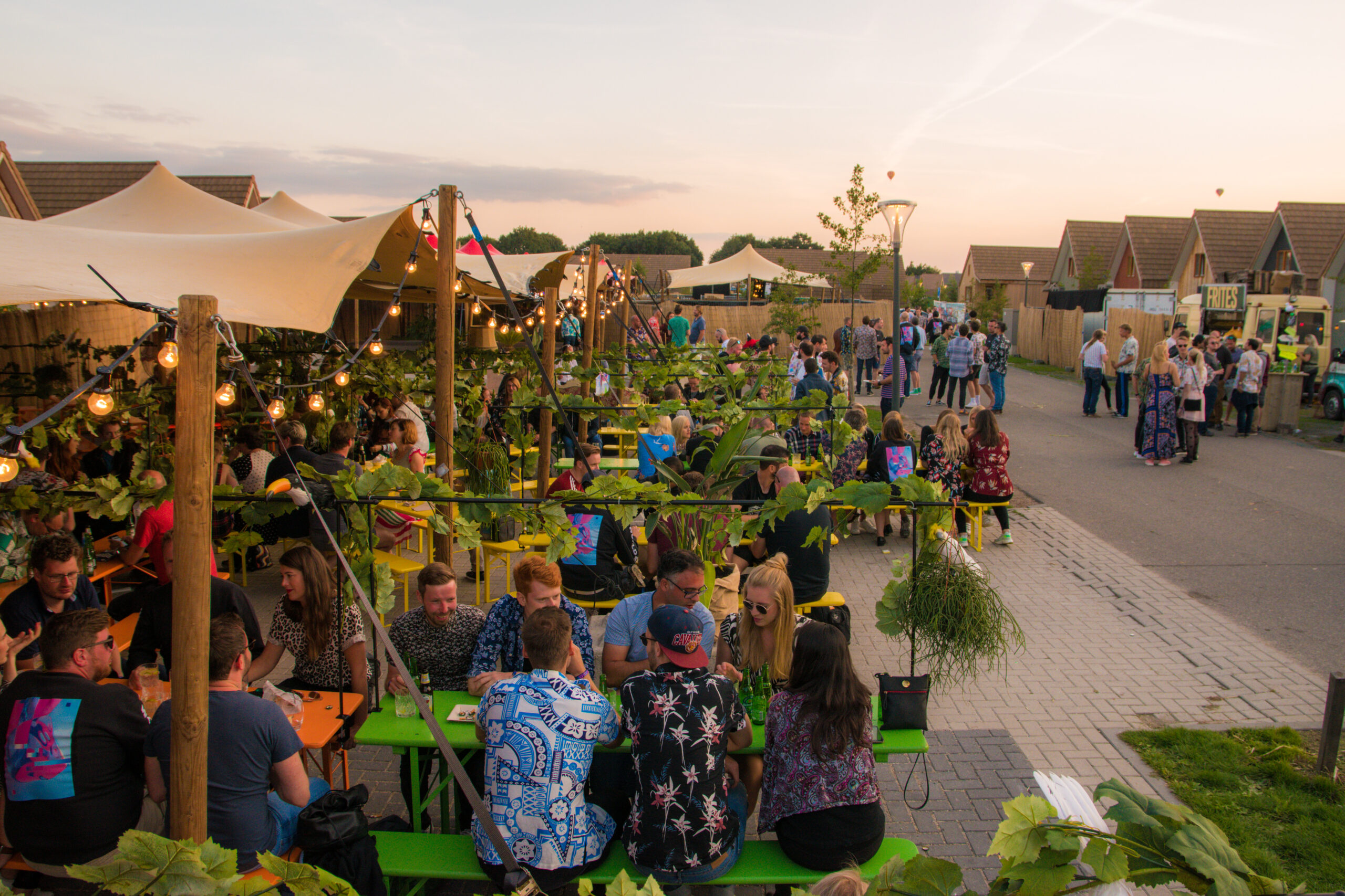Een sfeervol outdoor festivalterrein in de avondschemering, met verlichte stretchtenten, houten tafels en veel groen, waar bezoekers gezellig samen eten en drinken.